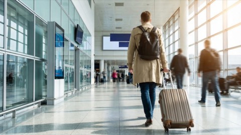 Man in beige trench coat pulling silver suitcase walking through bright modern airport terminal with glass walls
