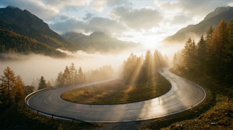 Mountain hairpin road at sunrise with misty valley golden sun rays wet tarmac low angle view and autumn trees foreground